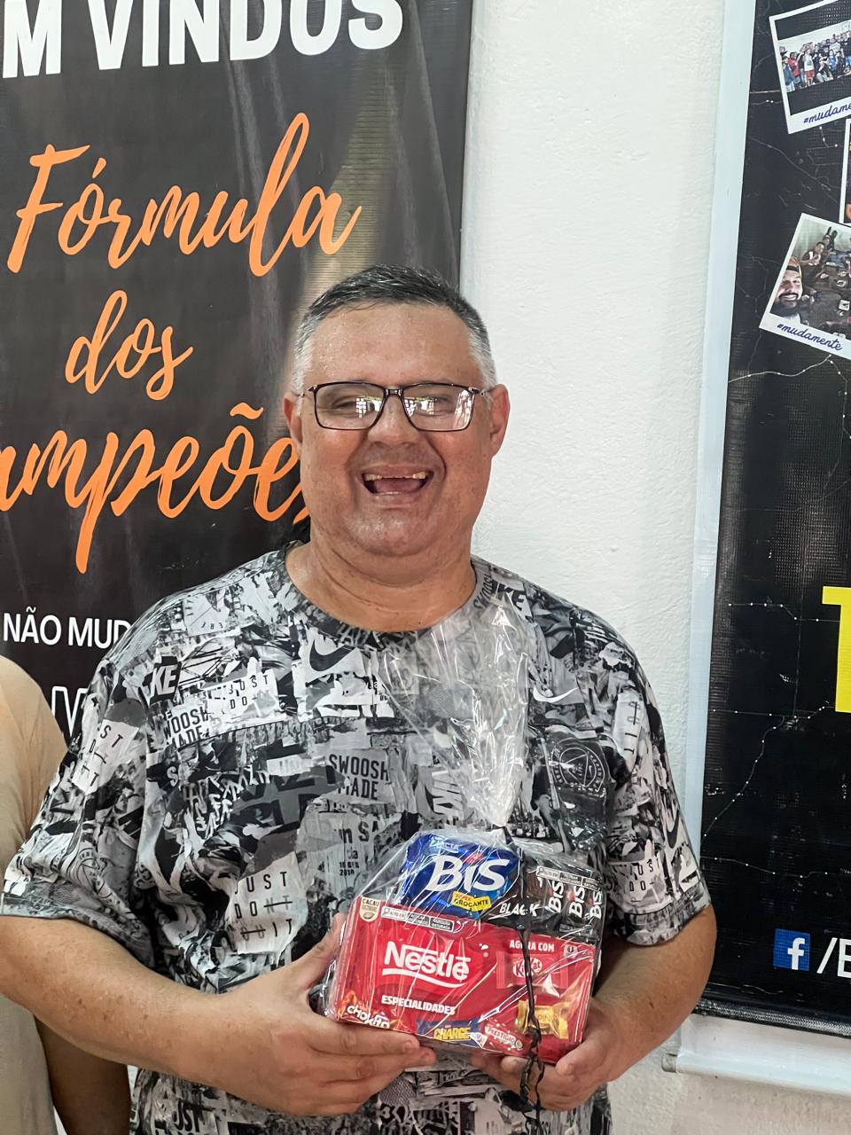 Homem sorrindo e segurando pequenos presentes diante de um painel.