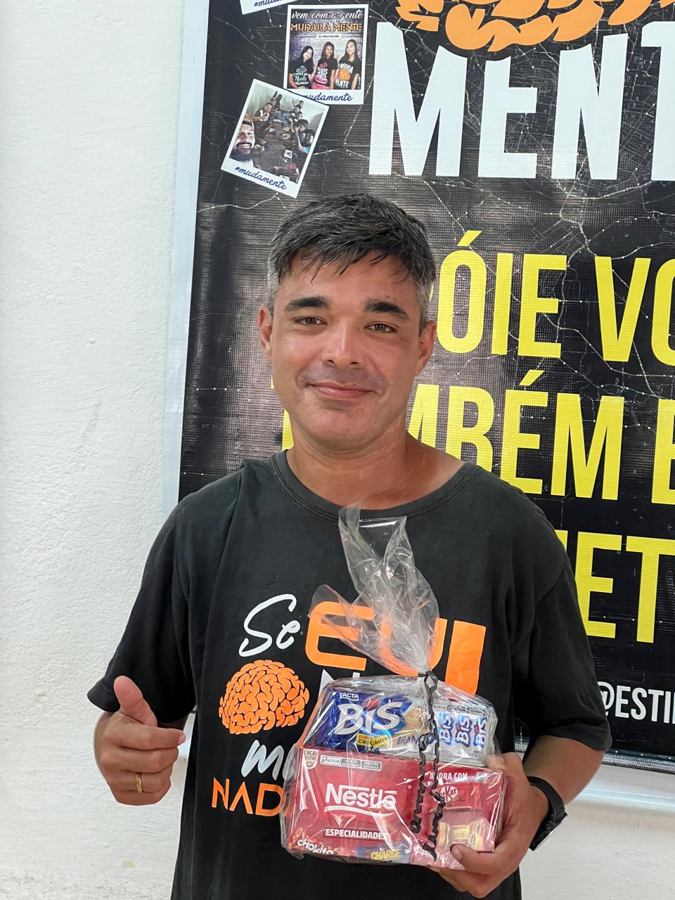 Homem segurando pequenos presentes e fazendo gesto positivo com a mão diante de um painel.