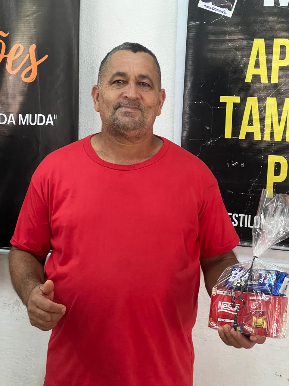 Homem de camiseta vermelha segurando pequenos presentes diante de um painel.